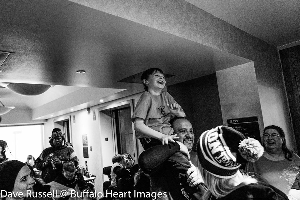 Black and white photo of young ADAPT activist Heath Montgomery being carried on the shoulders of personal care attendant Manny Corral. ADAPT activists rallied at the Housing and Urban Development offices in Washington, D.C. Infront of the group, are three activists holding up a blue shower curtain banner. ADAPT activists Nancy Salandra and one other activist sitting together. Group of ADAPT activists in blue shirts protesting in the capitol rotunda.