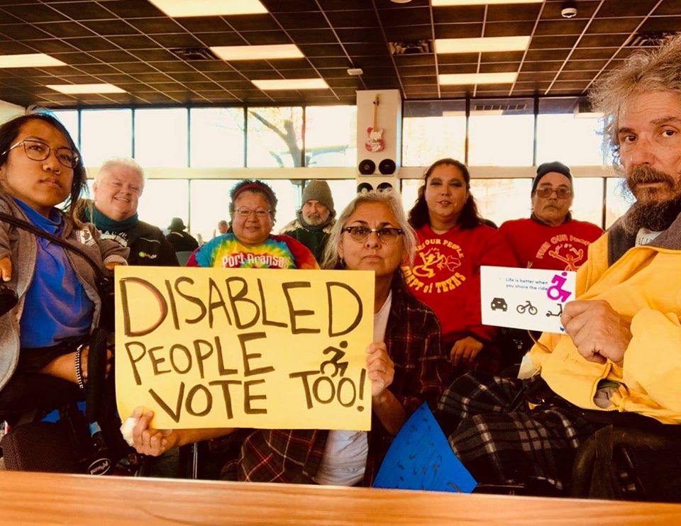 A group of ADAPT protesters stand inside of a building holding a sign that says "Disabled People Vote Too"