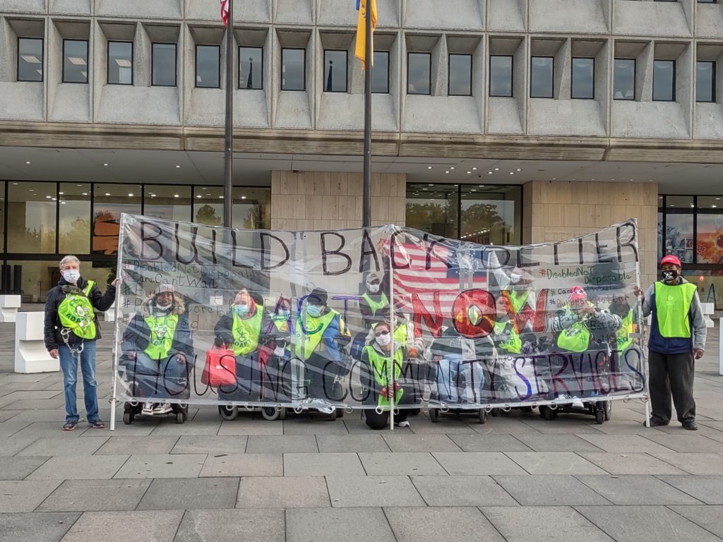 People wearing vests, protesting in front of a government building, holding a banner that reads: "Build Back Better Act Now"