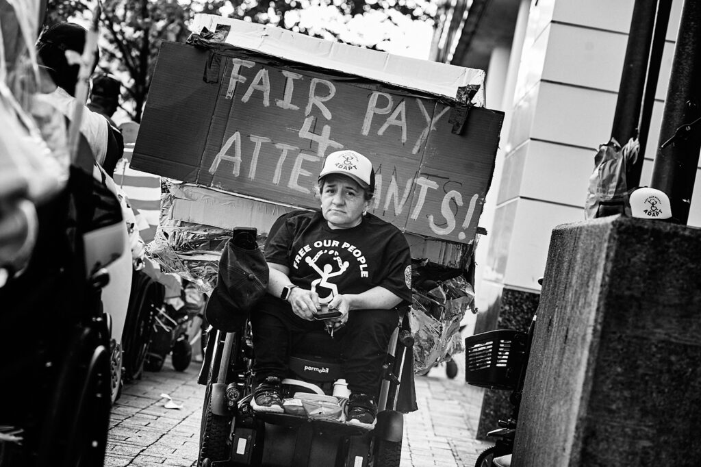 Person on Wheelchair with an ADAPT shirt. Banner behind the person reads: "Fair Pay 4 Attendands"