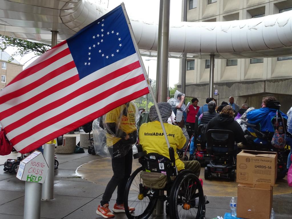 Person in wheelchair, with an american flag. Stars on flag have the shape of a person in a wheelchair.