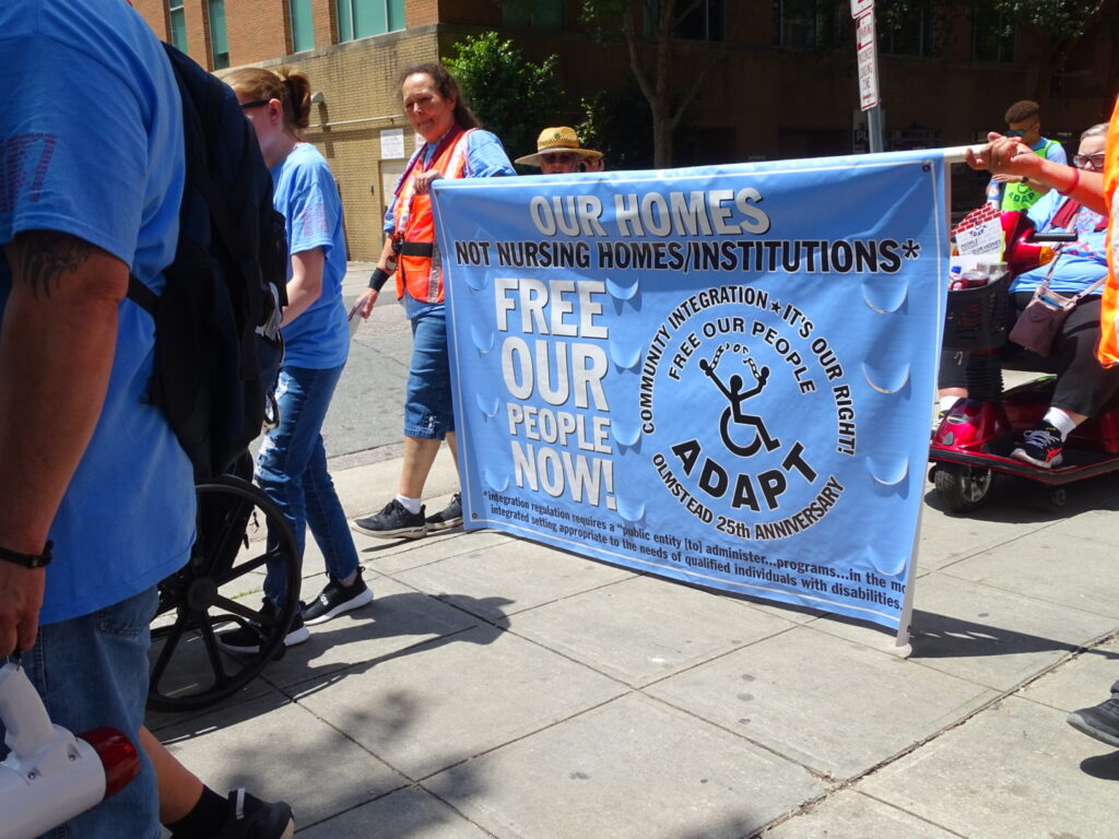 People marching, holding an ADAPT banner that reads: "Our homes, not nursing homes. Free our people now!"