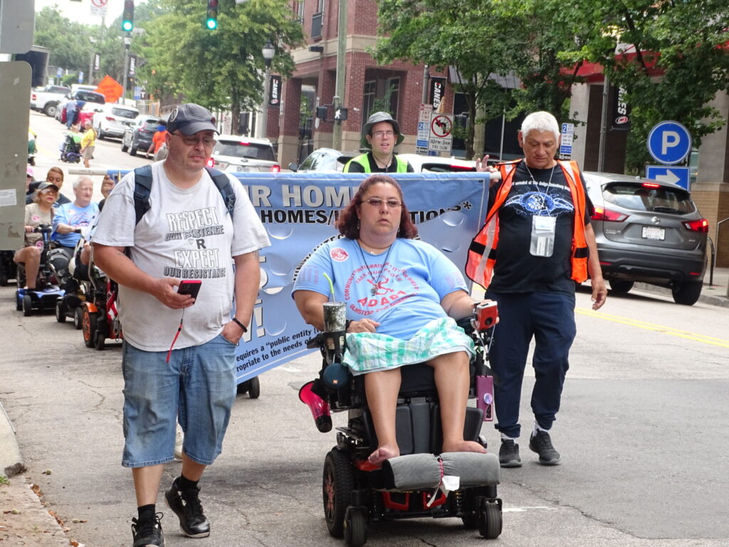 Woman on Wheelchair, marching next to two other people with a banner behind them.