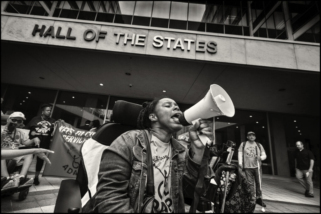 person wearing adapt shirt in wheelchair yelling into megaphone with others around